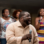 Man singing into microphone with three women backup singers