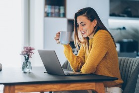 Woman drinking cup of coffee at table and smiling at laptop
