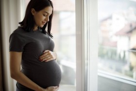 Pregnant woman holds stomach while standing next to a window.