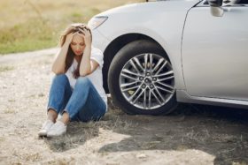 A woman who looks stressed sits on the ground next to a car.